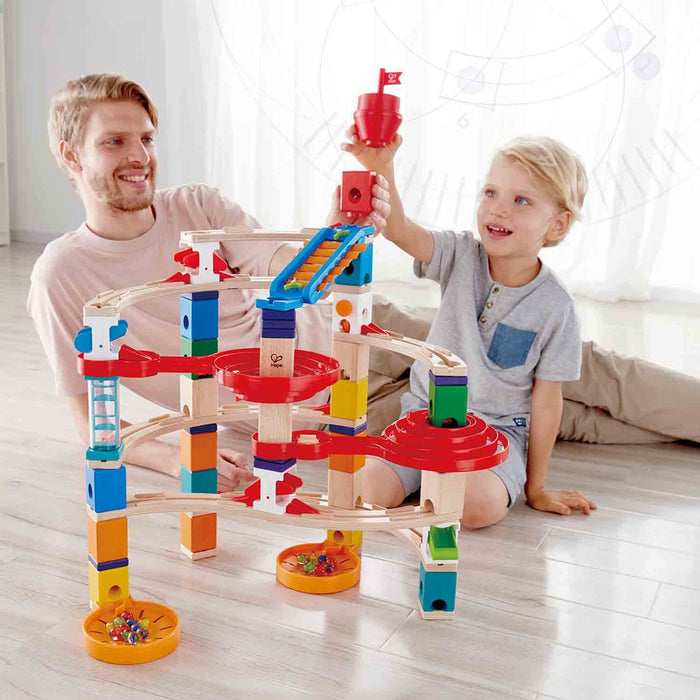 Father and son play with a wooden marble run featuring red spirals, funnels, and colorful blocks on a light wood floor.