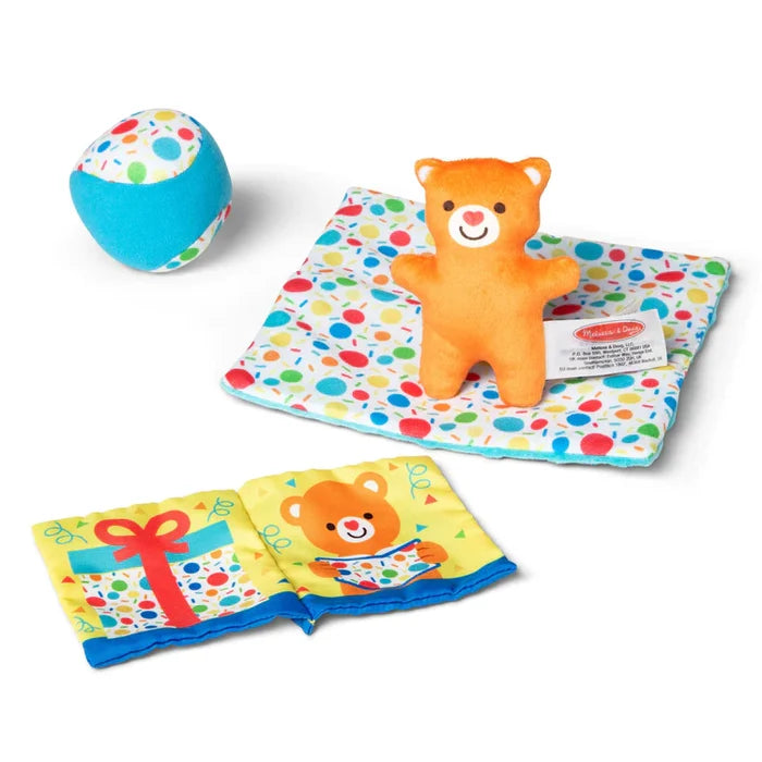 Plush orange bear on a polka dot mat, blue polka dot ball, and a cloth book with bear and gift illustrations.