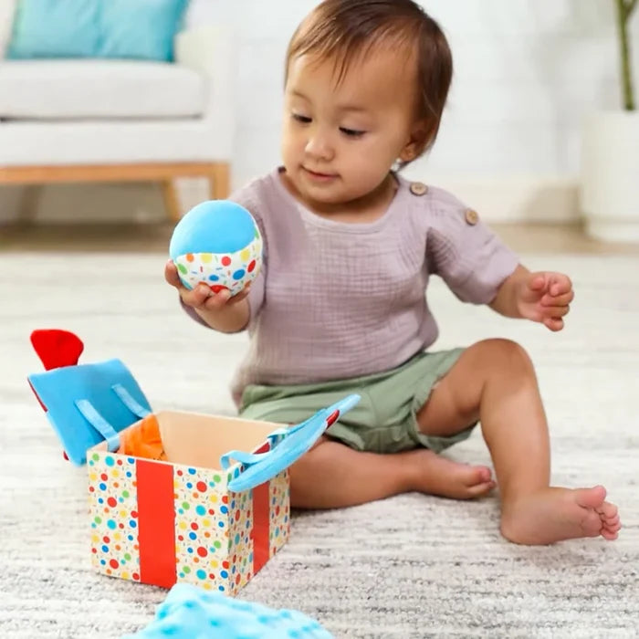 Toddler holding a blue soft ball while playing with an open wooden gift box toy featuring colorful polka dots.