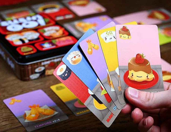 A hand holds a fan of colorful illustrated cards featuring cute food illustrations, with a matching game tin and additional cards blurred in the background on a wooden surface.