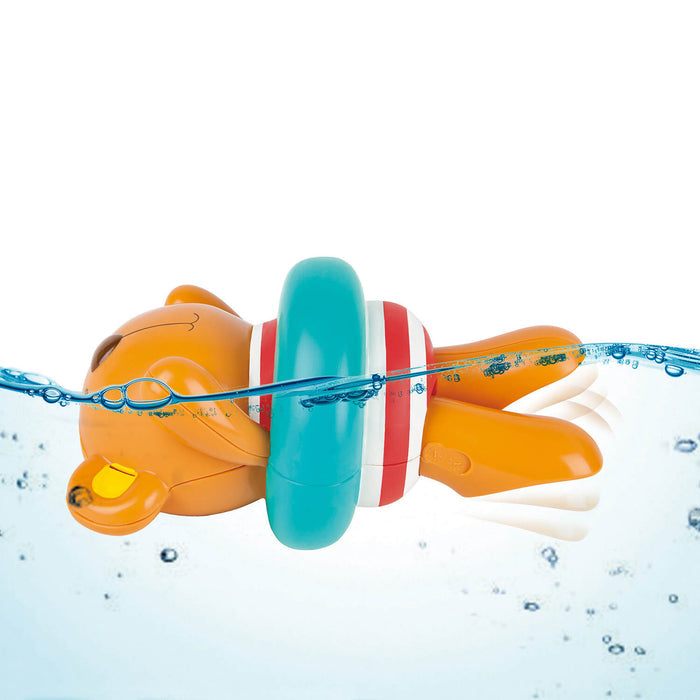 Orange bear toy wearing a teal ring and striped swimsuit, floating on its back in water with kicking legs.