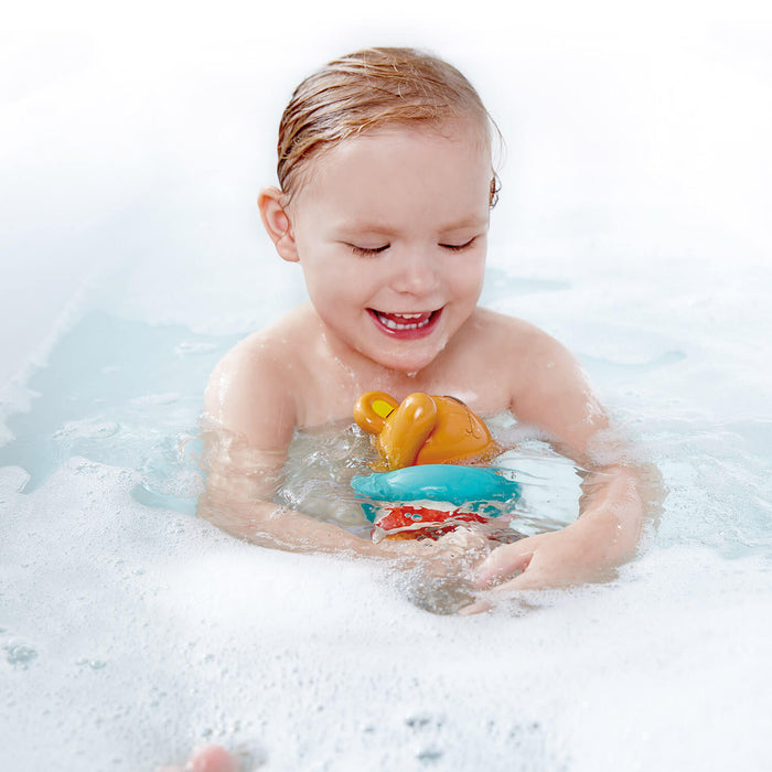 Smiling child in a bubble bath playing with an orange teddy bear toy in a blue and red inner tube.