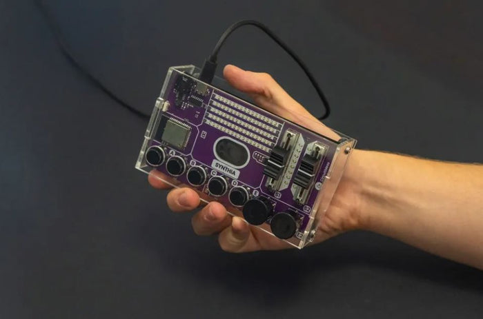 Hand holding a purple handheld synthesizer in a clear case with five black knobs, two sliders, and a screen.