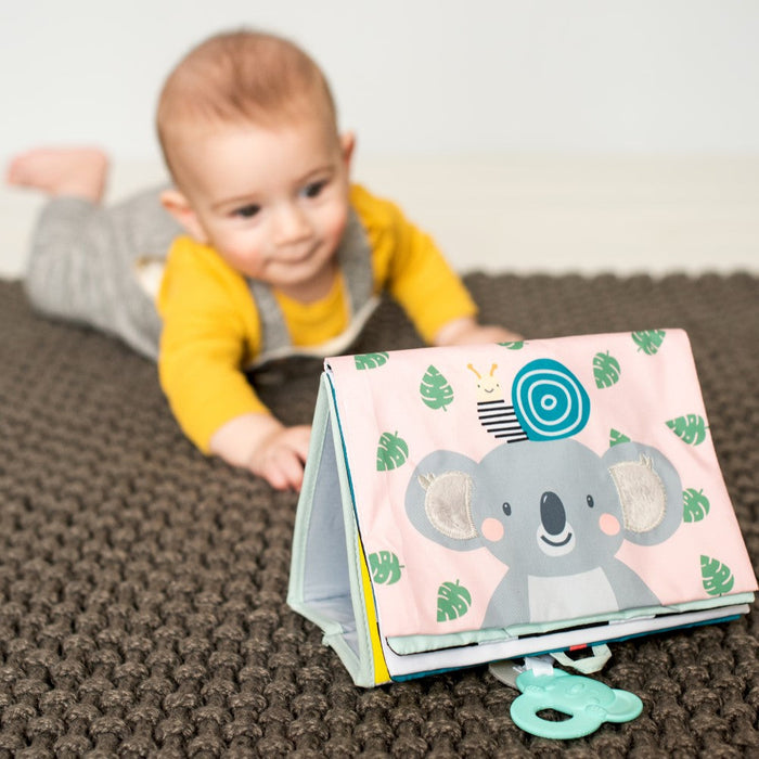 Baby on tummy time looks at a standing fabric book with a pink koala illustration and teal teether on a brown rug.