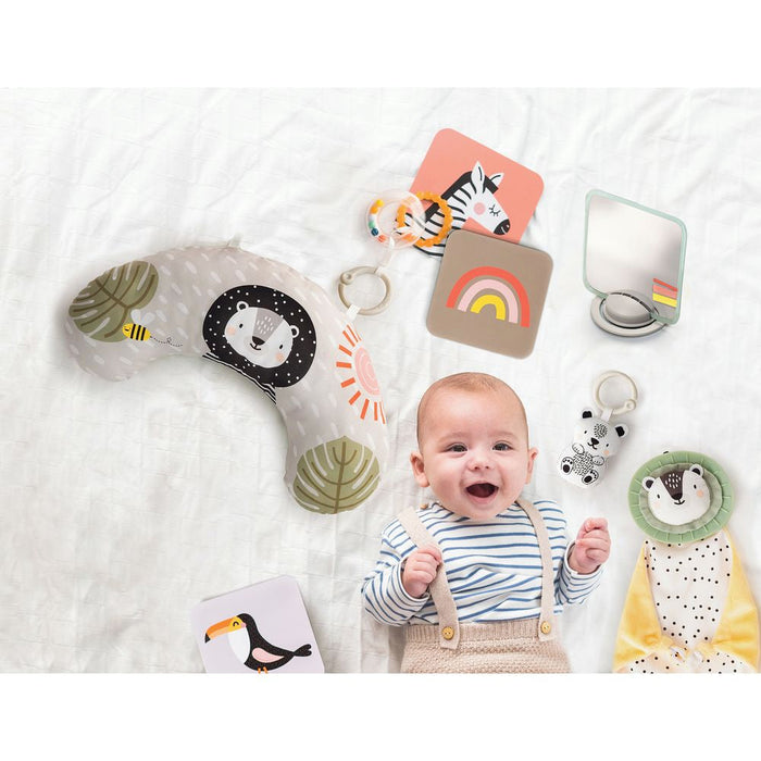 Smiling baby on a white sheet with a lion-patterned pillow, animal flashcards, mirror, and plush sensory toys.