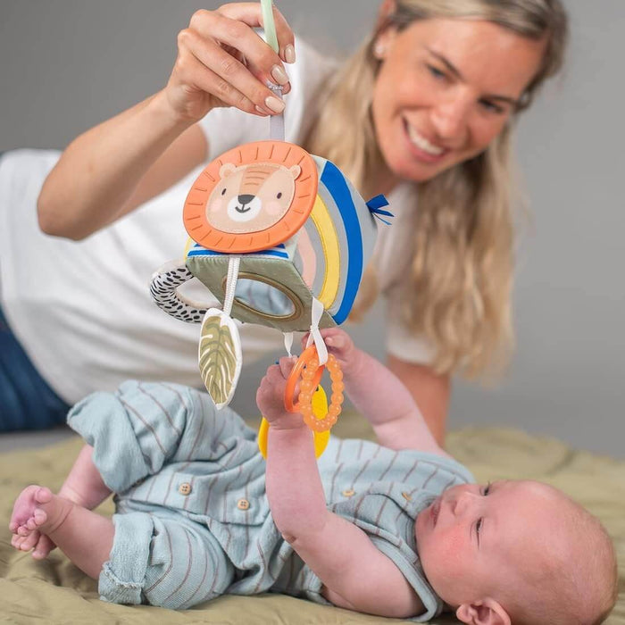 Woman holds a colorful lion sensory cube over a baby reaching for textured rings, a mirror, and a leaf.