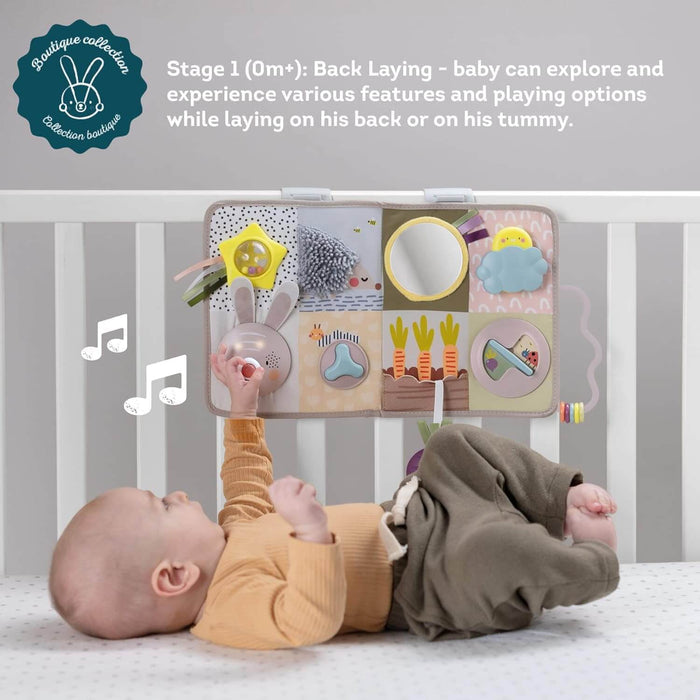 Baby in a crib reaches for a rabbit button on a sensory fabric activity board with a mirror, sun, and carrot toys.