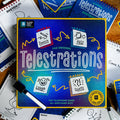 Blue Telestrations game box with yellow trim, surrounded by dry-erase markers and pads showing cartoon gameplay icons.