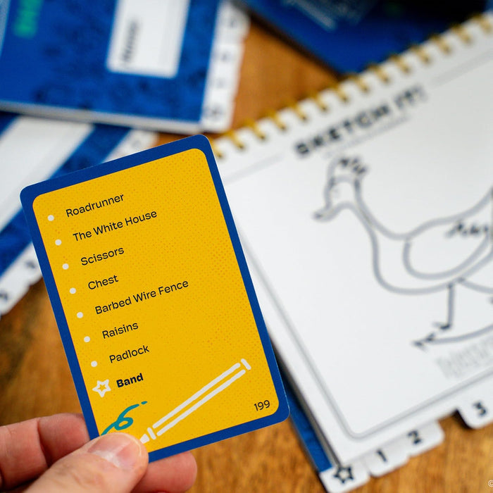 A hand holds a yellow game card with a word list over a spiral notebook featuring a simple line drawing of a bird.