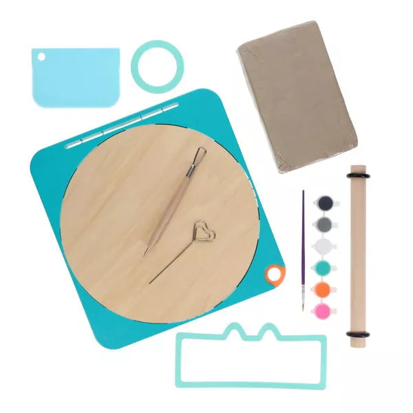 Pottery kit with teal base, wooden wheel, grey clay block, six paint pots, brush, rolling pin, and sculpting tools.