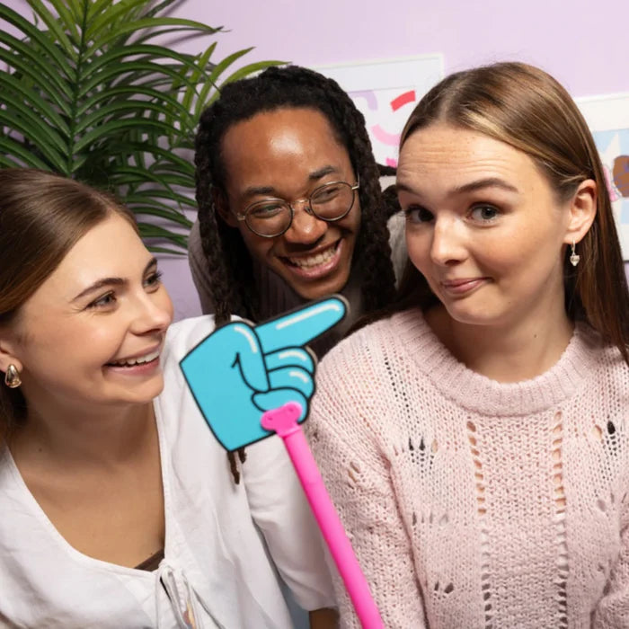 Three friends laughing as a large blue cartoon hand on a pink stick points toward one woman.