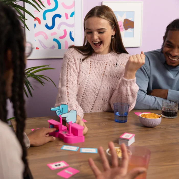 Friends playing a game with a pink voting device featuring blue pointing hands and cards on a wooden table.