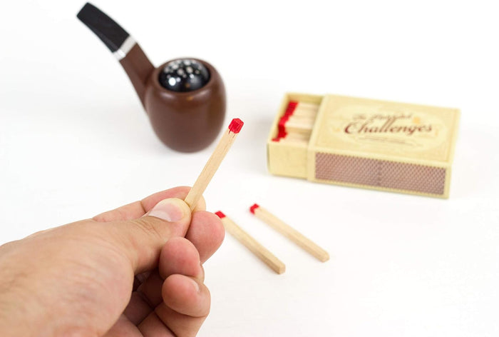 Hand holding a red-tipped matchstick next to a vintage matchbox and a brown toy smoking pipe on a white background.