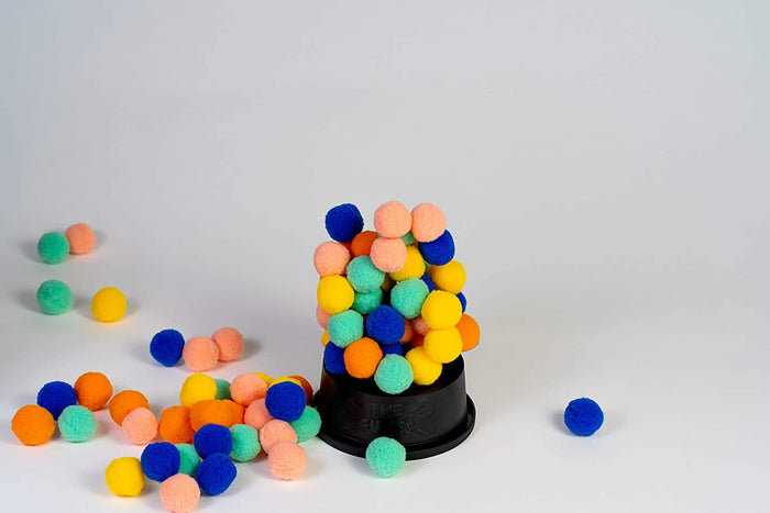 Colorful fuzzy pom-poms stacked in a tower on a black circular base with others scattered on a white background.