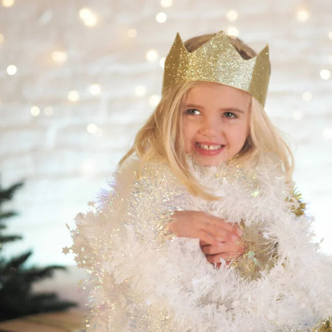 Gold glitter crown with points worn by a smiling girl wrapped in white tinsel against festive, glowing lights.