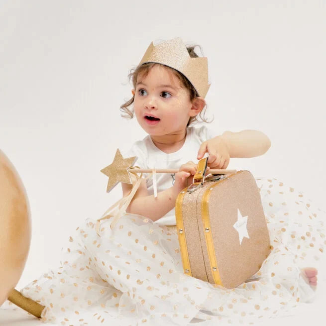 Toddler wearing a shimmering gold glitter crown with triangular peaks, holding a matching star wand and suitcase.