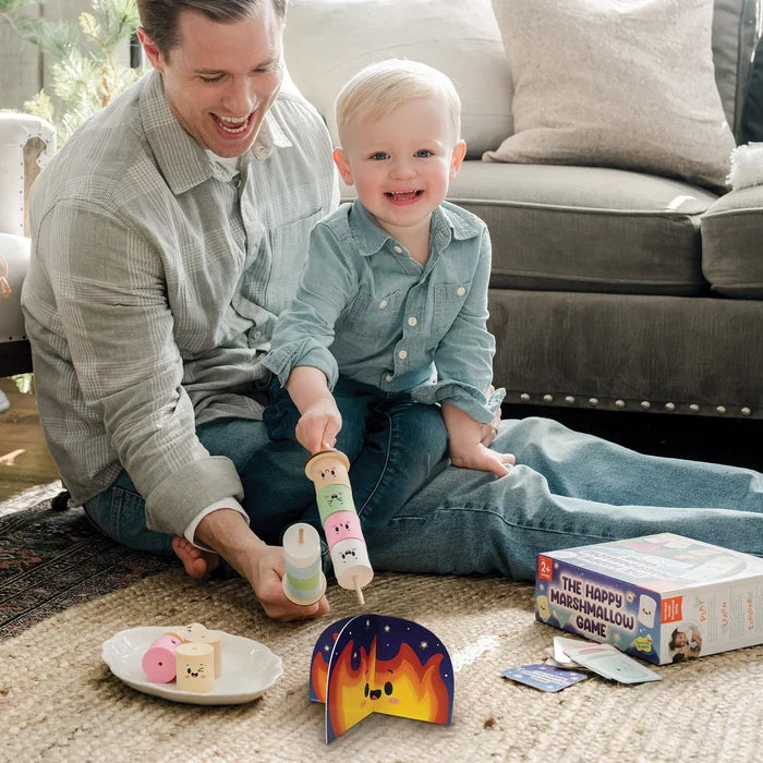 Father and son roast colorful wooden marshmallows with faces over a small cardboard campfire on a rug.