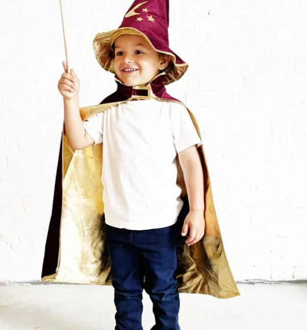 A child wears a bordeaux velvet wizard hat with gold stars and a matching gold-lined cape, holding a wooden wand.