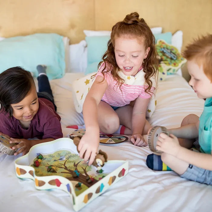 Three kids play a board game using squirrel-shaped tongs and colorful acorns on a tree-shaped tray on a white bed.