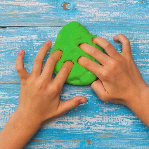 Hands pressing and stretching a lump of bright green therapy putty on a rustic blue wooden background.