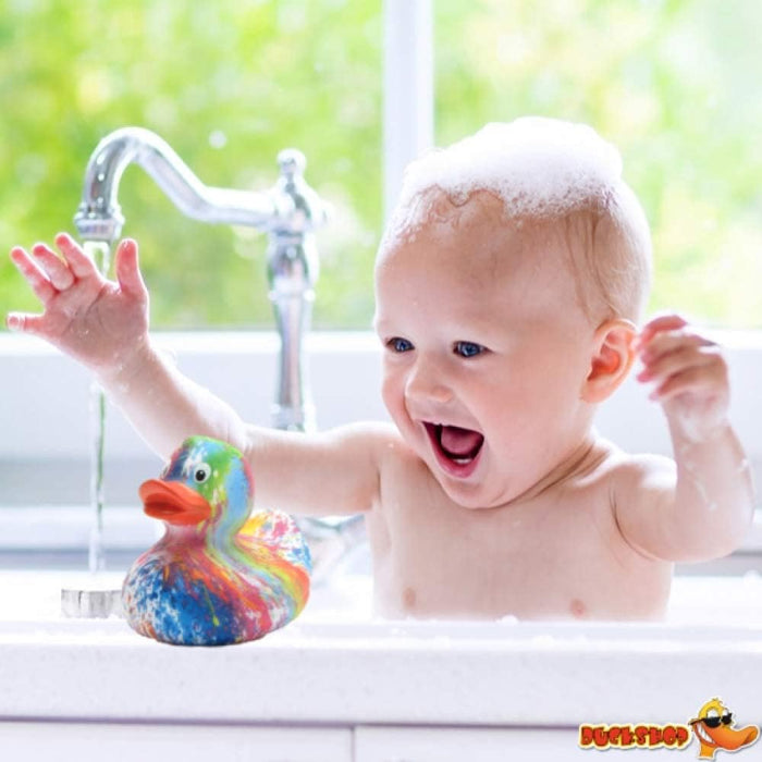 Rainbow swirled rubber duck with an orange beak on a white ledge next to a happy baby covered in soap bubbles.