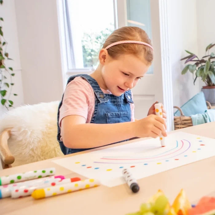 A girl draws a rainbow using a polka-dot paint marker on paper, with more colorful markers on a wooden table.