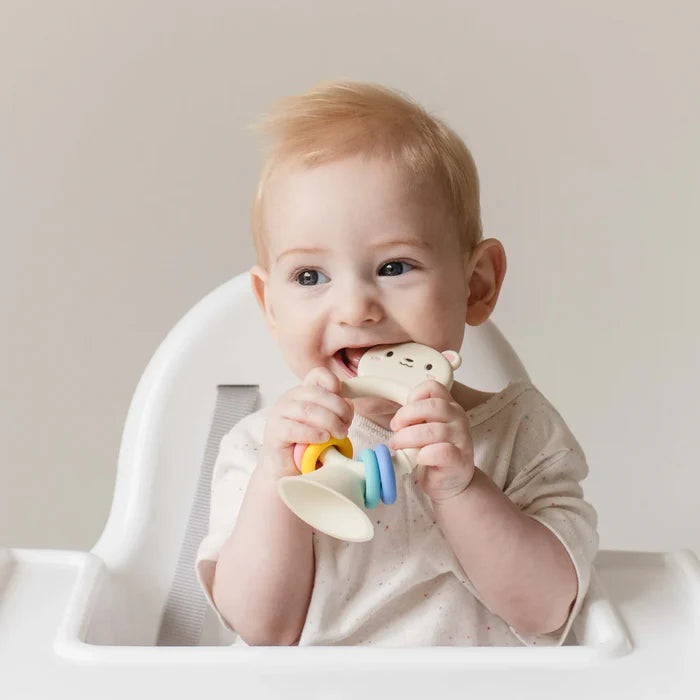 Baby in a high chair chewing a cream silicone bear teether with colorful rings and a suction cup base.