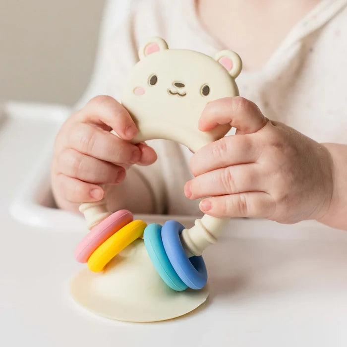 Baby holding a cream bear-shaped silicone toy with four colorful rings and a suction base on a high chair tray.