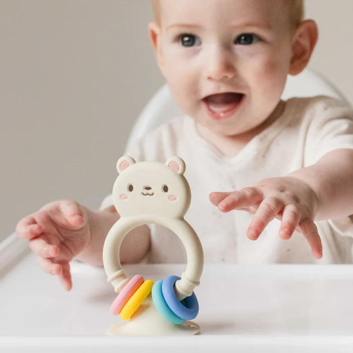 Cream silicone bear toy with colorful rings on a high chair tray, with a smiling baby reaching for it.