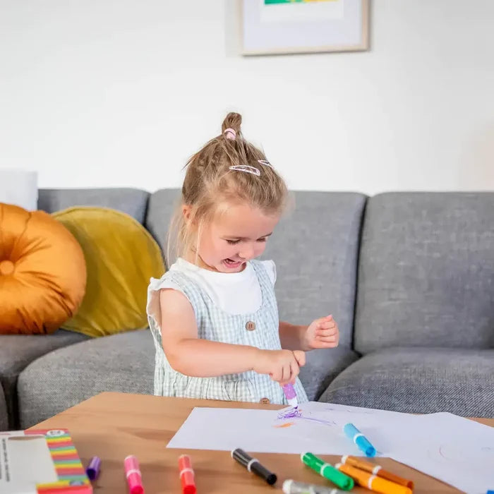 Young girl drawing on white paper with a purple marker, surrounded by colorful chunky markers on a wooden table.
