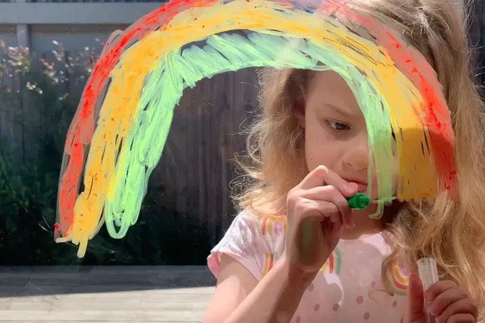 A girl draws a vibrant rainbow on a glass window with a green silk crayon, showing smooth, paint-like strokes.