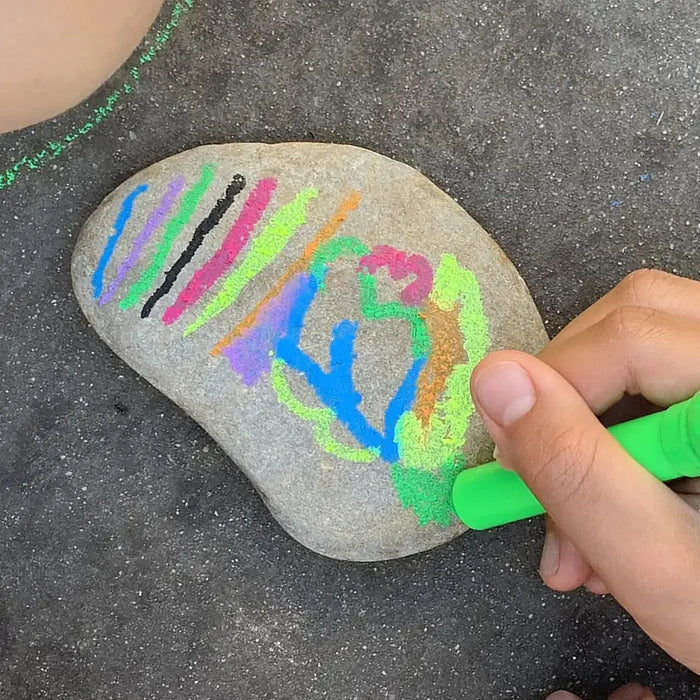 A hand draws on a smooth rock with a neon green silk crayon, showcasing vibrant streaks of pink, blue, and yellow.