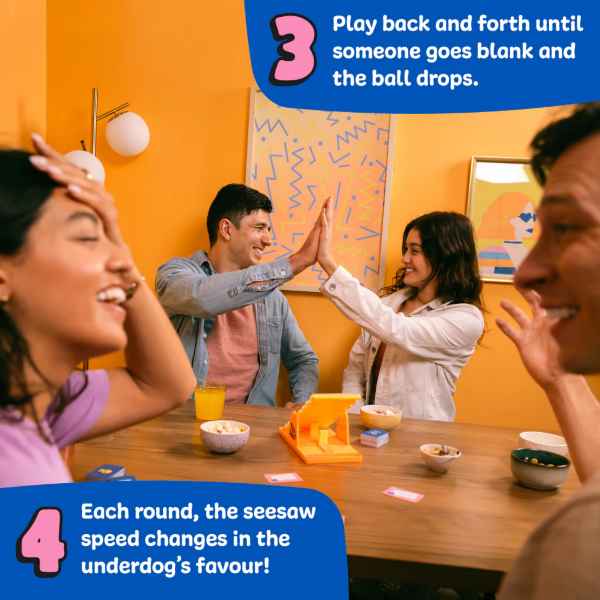 Friends high-fiving and laughing while playing a game with a yellow seesaw on a table against orange walls.