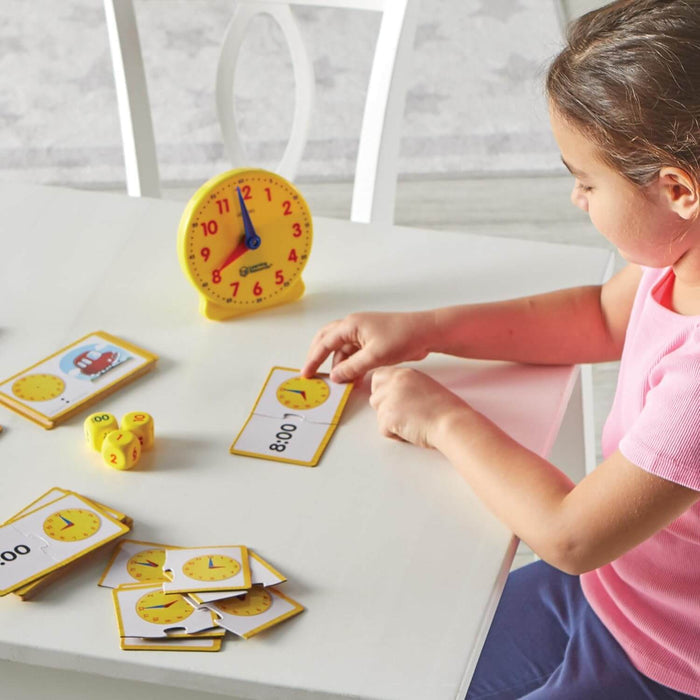 A child matches puzzle cards showing 8:00 next to a yellow analog toy clock and numbered dice on a white table.