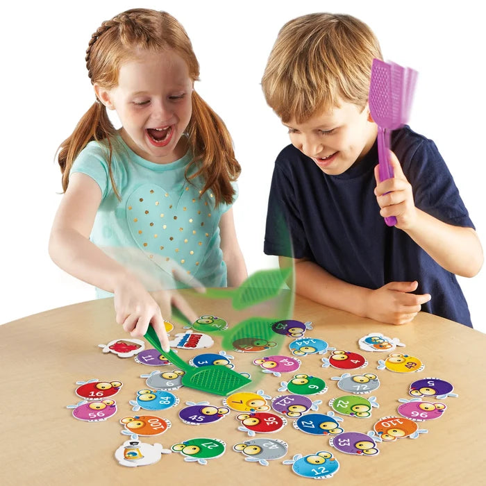 Two children use plastic swatters to hit colorful numbered bug cards spread across a table.