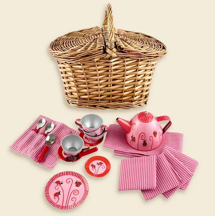 Wicker basket with a pink and red ladybug-themed tin teapot, cups, plates, and striped napkins.