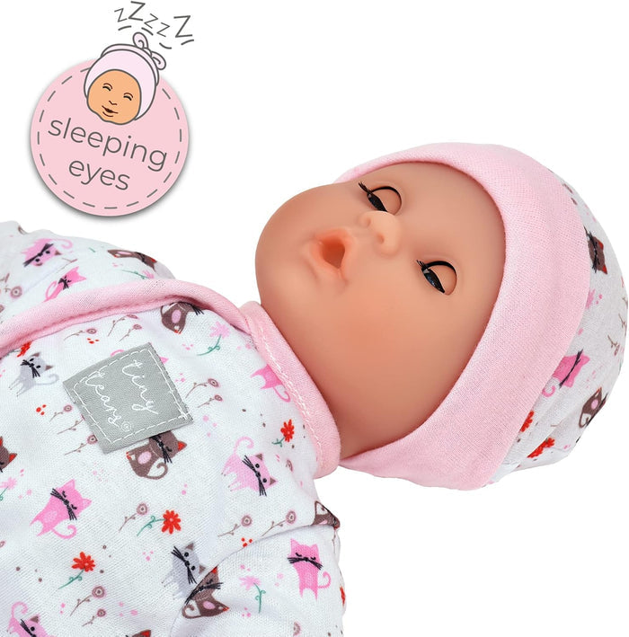 Baby doll with half-closed sleeping eyes wearing a pink cat-patterned onesie and matching hat.