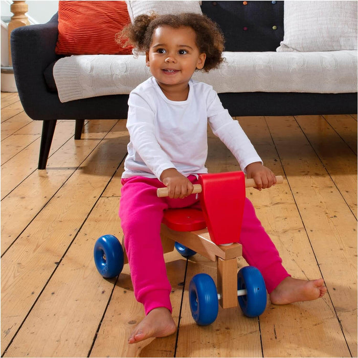 Toddler on a wooden ride-on trike with four blue wheels and red accents on a hardwood floor.