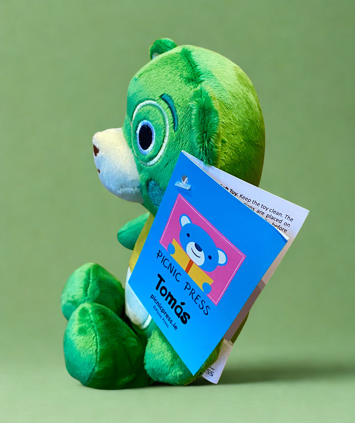 Profile of a green plush teddy bear with a cream muzzle, blue eyes, and a blue tag labeled Tomás by Picnic Press.