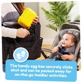 Adult placing a yellow plastic egg box into a black travel bag with a smiling baby in a stroller nearby.