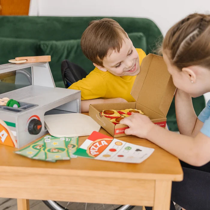 Two kids playing with a wooden pizza counter set including an oven, pizza box, toppings, play money, and menu.