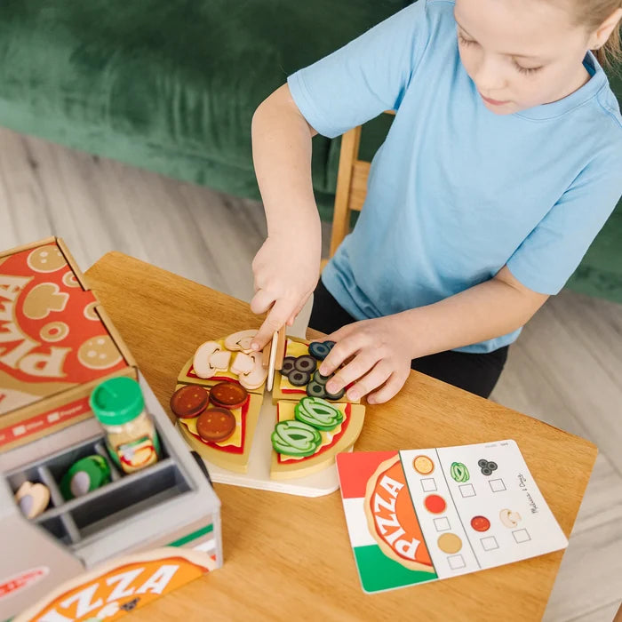 A child slices a wooden toy pizza with colorful toppings next to a play counter and menu card on a wooden table.