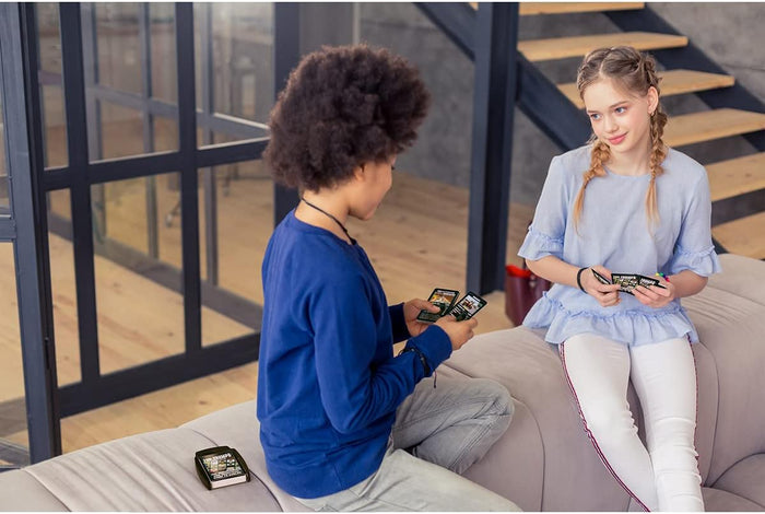 Two children playing with black Roblox Top Trumps guide cards. A black plastic storage case sits on the grey sofa.