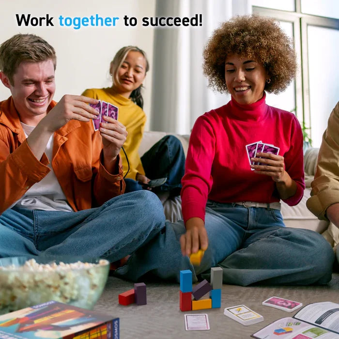 Four friends smile and play a cooperative game, stacking colorful blocks and holding cards on a living room floor.