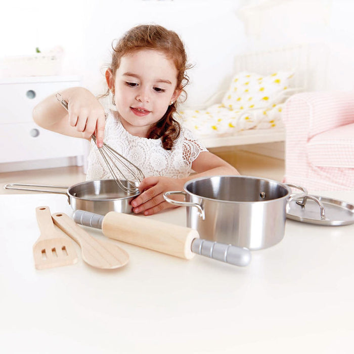 Young girl playing with stainless steel toy pots, a metal whisk, and wooden kitchen utensils on a white table.