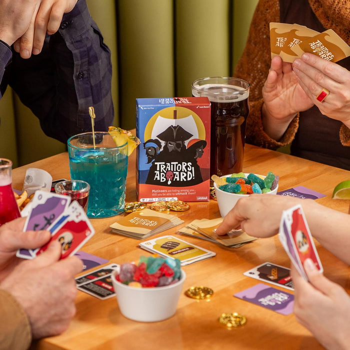 People play Traitors Aboard card game with a colorful box, gold coins, and cocktails on a wooden table.