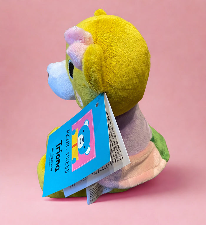 Yellow plush teddy bear from the side with a blue Tríona tag and pink inner ear, against a pink background.