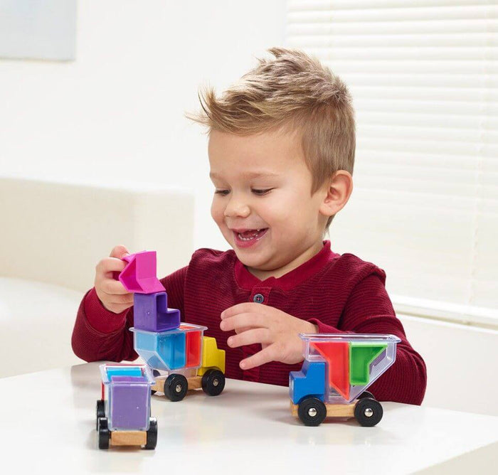 Child loading colorful geometric blocks into three wooden toy trucks with clear containers on a white table.