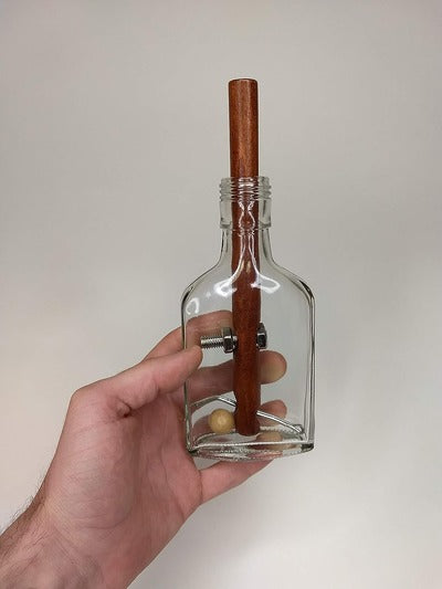 Hand holding a clear glass bottle puzzle containing a wooden rod, a metal bolt with a nut, and a wooden ball.