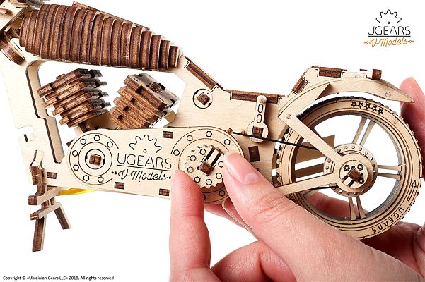 Hands demonstrating the mechanical belt drive and gears on a light brown laser-cut wooden motorcycle model.